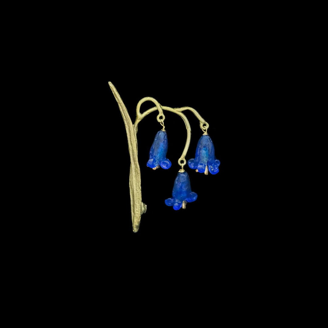 Eine Brosche im Vintage-Stil in Grüntönen mit drei baumelnden blauen glockenförmigen Glasblüten an einem geschwungenen Stiel vor schwarzem Hintergrund.