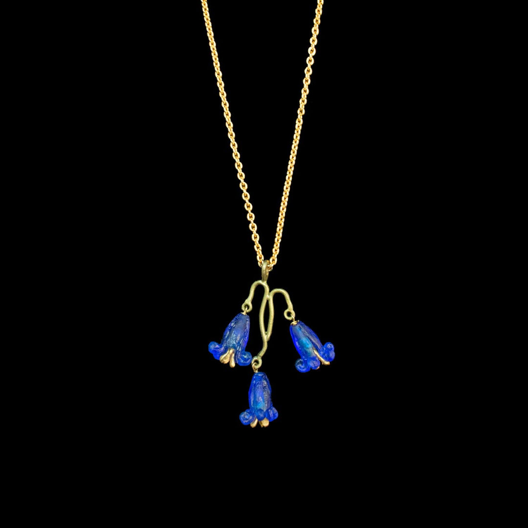 Eine goldene Halskette mit drei baumelnden, glockenförmigen blauen Blüten mit goldfarbenen Konturen vor schwarzem Hintergrund.